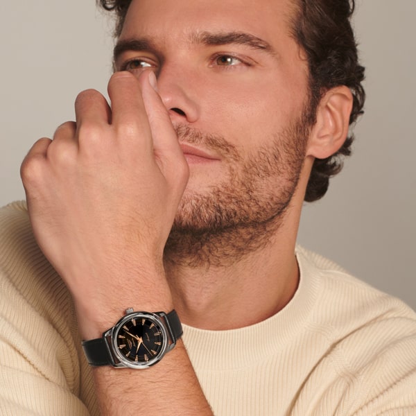Ein Mann mit kurzem, lockigem braunem Haar und gestutztem Bart, der einen cremefarbenen Pullover und eine Longines Heritage Classic-Armbanduhr trägt, legt seine Hand vor sein Gesicht und blickt nachdenklich in die Ferne.